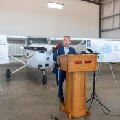 Un homme en costume parle derrière un podium dans un hangar d'avion. On peut voir un petit avion et des affiches en arrière-plan.