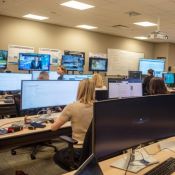 Bureau à aires ouvertes où de nombreuses personnes travaillent derrière des écrans d'ordinateur affichant des reportages d'actualité.
