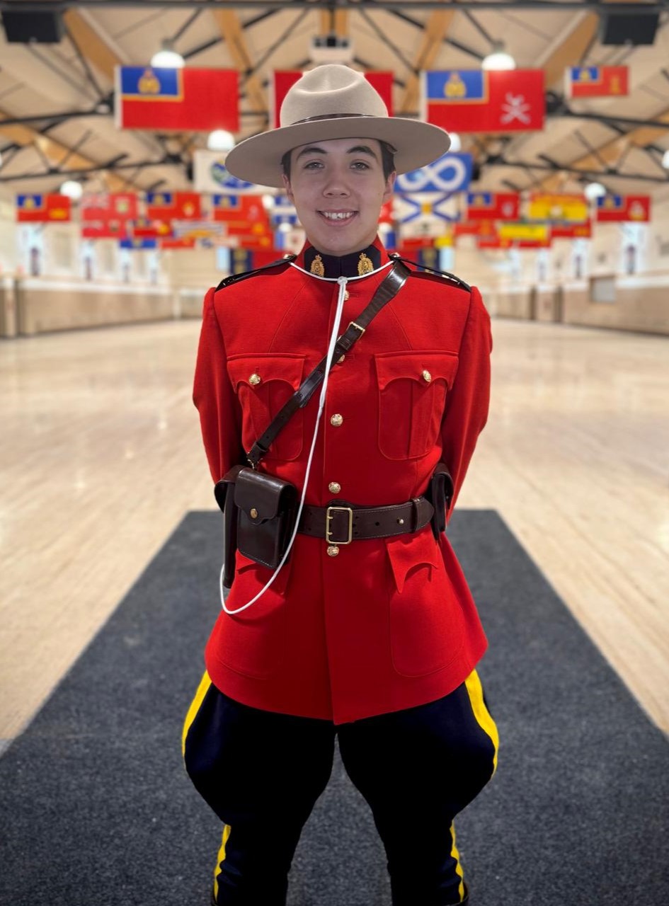 Un policier en tunique rouge est debout dans un gymnase et on aperçoit des drapeaux provinciaux du Canada suspendus au plafond à une certaine distance derrière lui.