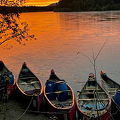 Cinq canots sont accostés en bordure d'une rivière. Un coucher de soleil d'un orange rougeâtre se reflète sur les eaux calmes. On aperçoit les montagnes en arrière-plan. 