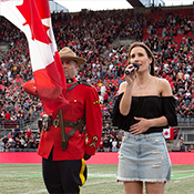 Un agent de la GRC portant le drapeau du Canada et une chanteuse tenant un micro se tiennent debout, côte à côte, sur un terrain de football devant la foule réunie dans le stade.