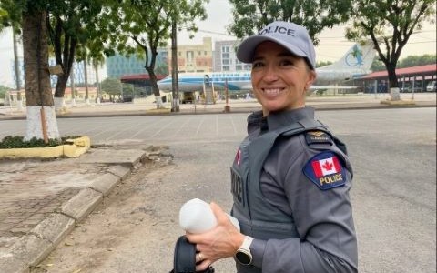 Une femme en uniforme de police sourit dans un stationnement avec un bâtiment et un avion au loin.