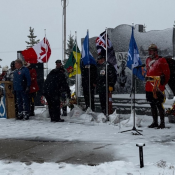Un agent de la GRC en tunique rouge salue un certain nombre de drapeaux devant un monument commémoratif enneigé.