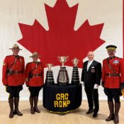 Trois policiers de la GRC et le gardien officiel de la Coupe Grey posant debout avec la Coupe Grey, la Coupe Connaught et la Coupe Minto devant un immense drapeau du Canada.