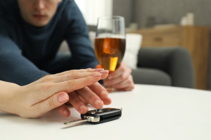 Gros plan d'une clé de voiture sur une table en avant-plan; une personne est assise en arrière-plan et elle tient un verre contenant une boisson alcoolisée. 