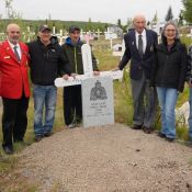 Neuf personnes debout autour d’une tombe marquée d’un monument de la GRC et d’une croix au milieu d’un cimetière rempli de croix.