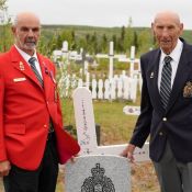 Deux hommes âgés en veston à côté d’un monument régimentaire de la GRC dans un cimetière rempli de croix.
