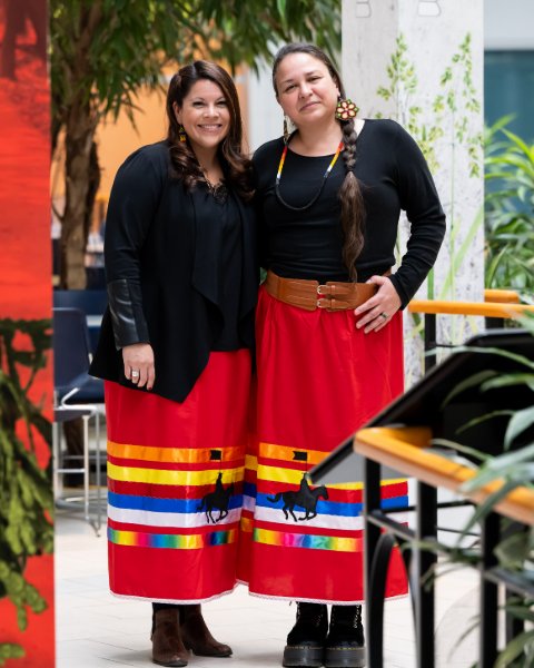 Deux femmes vêtues d’un chandail noir et d’une jupe longue rouge au motif de l’emblématique gendarme à cheval de la GRC et de rubans multicolores sourient.