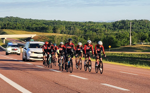 On aperçoit plusieurs cyclistes le long de la route. Un véhicule de la GRC suit derrière eux.
