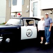 Une femme âgée et un homme posent à côté d’une voiture de police d’époque.