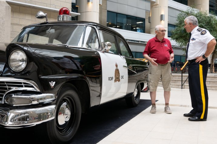 Un officier de la GRC en uniforme et un homme âgé vêtu d’un chandail rouge se tiennent à côté d’une voiture d’époque dans le hall d’entrée d’un immeuble.