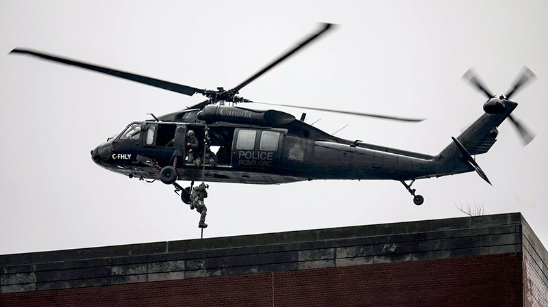 Un hélicoptère Black Hawk UH-60 de la GRC survole le toit d’un immeuble et une personne sort de l’hélicoptère à l’aide d’une corde pour se poser sur le toit.