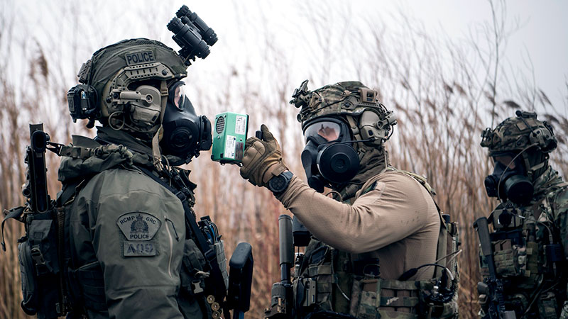 Trois personnes en tenue tactique utilisent des outils de protection contre des produits chimiques, biologiques, radiologiques, nucléaires sur un policier de la GRC. 