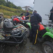 Deux membres de la GRC en uniforme préparent leur équipement pour partir en patrouille. L’un d’eux verse de l’essence dans un VTT vert avec un bidon d’essence.