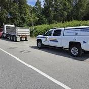 Un gros camion à benne accroché à une dépanneuse rouge sur le bord de la route, et une camionnette d’inspection des véhicules commerciaux de la police stationnée derrière lui.