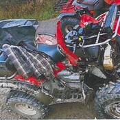 Un VTT rouge et noir, dont l’avant est fortement endommagé, se trouve sur l’accotement en gravier d’une route, avec un deuxième VTT rouge visible à l’arrière-plan.