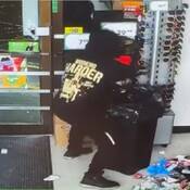 On voit un individu, dos à la caméra, tenir un objet au-dessus d'une grande poubelle. La personne est vêtue de noir et porte un manteau ou un chandail à capuchon noir avec un grand logo jaune dans le dos avec les mots « WORKING HARDER ».