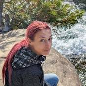 Photo d’Ashley Bosma, une femme de 40 ans vraisemblablement de race blanche, qui est accroupie sur un rocher près d’un cours d’eau tumultueux et qui regarde dans l’objectif de l’appareil photo. Elle a de longs cheveux rouge vif attachés en queue de cheval et elle porte un chandail muni d’un capuchon arborant un motif de losanges.