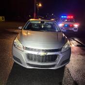 Une voiture Chevrolet grise est visible sur le bord de la route, avec un VUS de police derrière elle. Les gyrophares rouges et bleus du véhicule de police sont allumés.