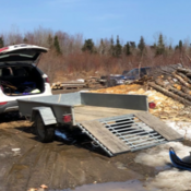 Une remorque utilitaire en aluminium, avec sa rampe abaissée, est attachée à un VUS blanc. Le hayon du VUS est ouvert et la remorque est vide.