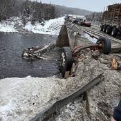 Remorque-citerne endommagée dans une rivière sous un tablier de pont gravement endommagé et un semi-remorque endommagé parmi des débris sur le tablier du pont.