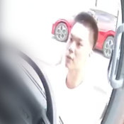 Un homme d’origine asiatique dans la vingtaine, qui porte un tee-shirt blanc et qui regarde dans un véhicule. Il y a une voiture rouge en arrière-plan.
