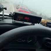 Une voiture rouge en bordure de route et, au premier plan, un radar de police affichant une vitesse de 106 km/h.