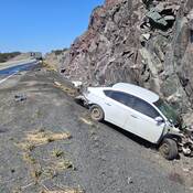 Une voiture blanche très endommagée adossée à une paroi rocheuse dans le fossé et, à l’arrière-plan, des camions de transport et d'autres véhicules.