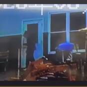 Un inconnu devant une porte d'entrée portant des vêtements foncés et amples, dont un chandail à capuchon avec le capuchon relevé. On aperçoit aussi de la machinerie lourde au premier plan de la photo.