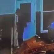 Un inconnu devant une porte d'entrée portant des vêtements foncés et amples, dont un chandail à capuchon avec le capuchon relevé. On aperçoit aussi de la machinerie lourde au premier plan de la photo.