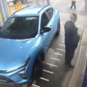 Un homme près de la portière côté conducteur d’un véhicule bleu, dos à la caméra de surveillance, portant des vêtements foncés.