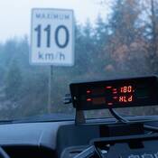 Un lecteur radar affiche 180 sur le tableau de bord d’une voiture de la PRCB. La voiture de patrouille se trouve sous la pluie devant un panneau de limite de vitesse qui indique 110 km/h
