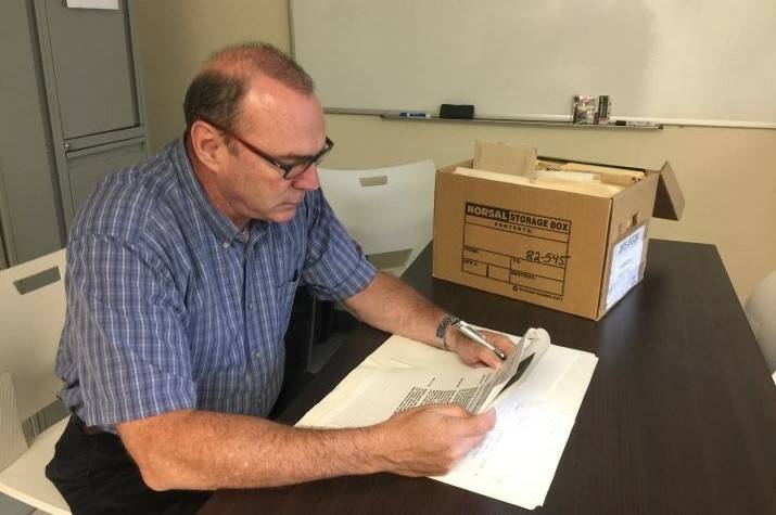 A male RCMP officer in civilian clothes looks over some papers, with a cardboard box on the table next to him. 