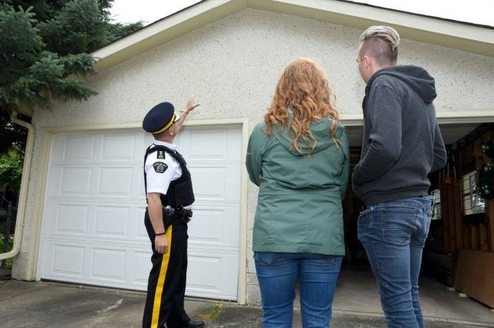 Male RCMP officer talks to a man and a woman in front of a garage.