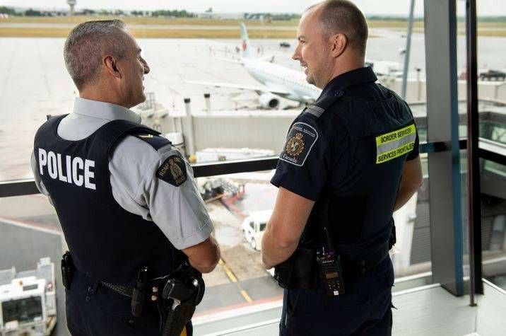 A male RCMP officer and a male CBSA agent talk to each other inside an airport. An airplane can be seen on the tarmac behind them. 