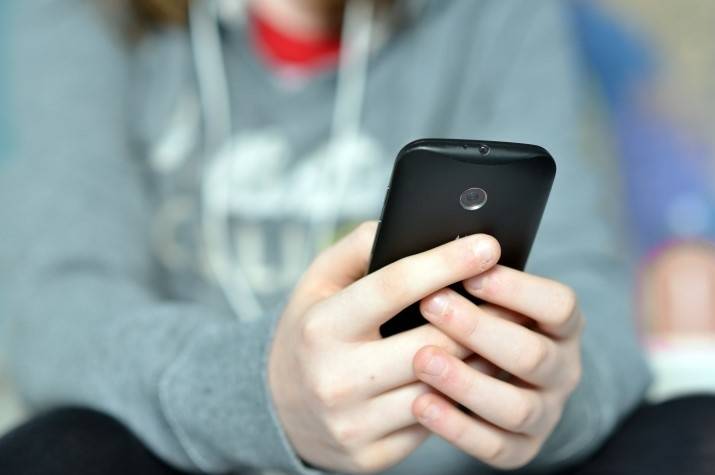 A close-up of hands that appear to belong to a teenage boy in a sweatshirt holding a cellphone.