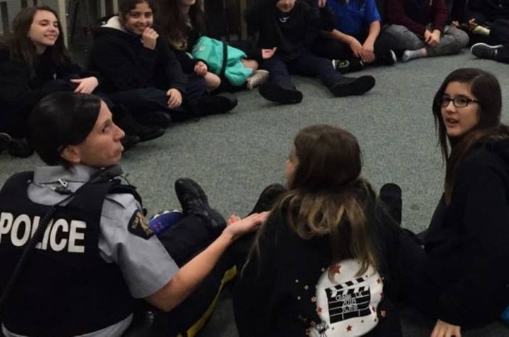 Female police officer sits in a circle on the floor of a classroom with young female students.