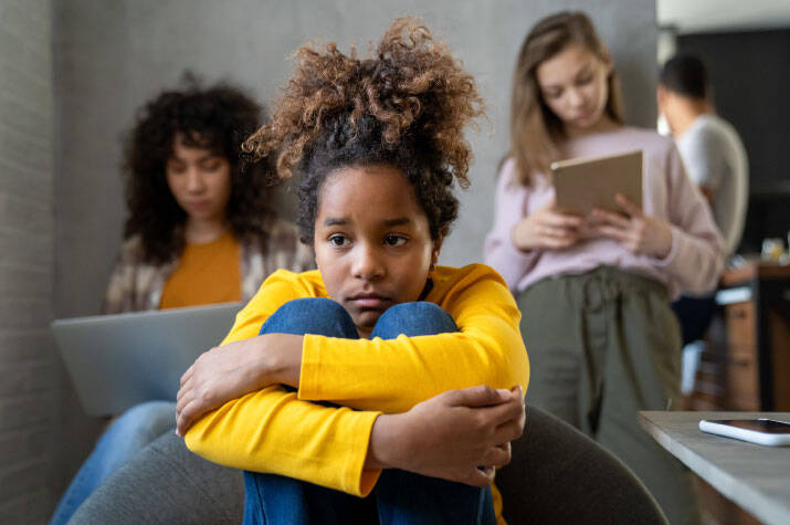 Child in a yellow long-sleeve shirt sits curled up in a chair with arms wrapped around bent knees. In the background, one person is using a laptop and another is using a tablet in an indoor living space.