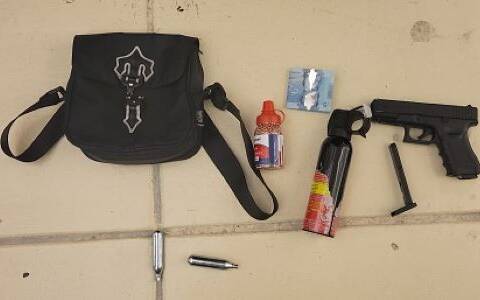 A black satchel, a five-dollar bill, a canister of bear spray and a black pellet hand gun laying on a tile yellow floor.