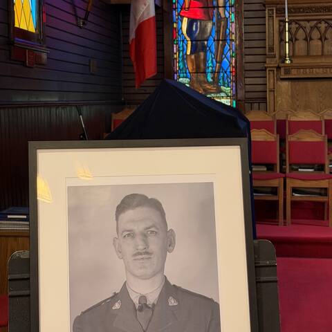 A black and white image of an RCMP officer sits in a frame in front of a stained-glass window inside a chapel