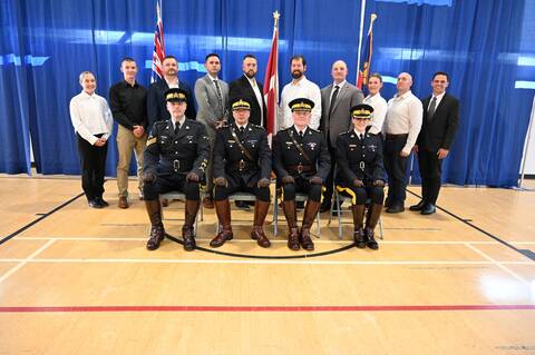 A group of 10 men and women wearing business attire standing behind a row of seated RCMP police officers, wearing ceremonial blue uniforms.