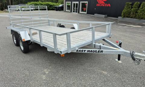 An aluminum/silver tandem axel utility trailer, with a wooden deck is parked in a lot. The words “EASY HAULER” are om black lettering on the side of the trailer’s tongue.