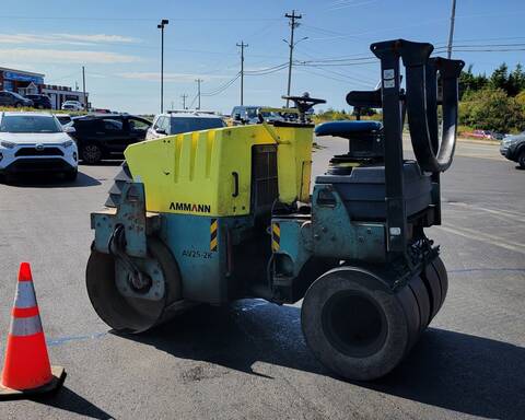 A yellow and green Ammann steamroller, viewed from the side, is stopped next to an orange traffic pylon in a parking lot. No one is on the steamroller and vehicles are visible in the background.