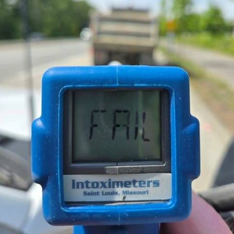 A blue roadside screening device with the word FAIL on it with a blurry dump truck in the background, parked on the side of the road.