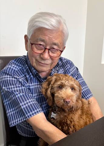 Man with grey hair, red glasses, blue shirt, holding a brown dog