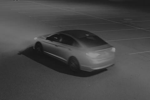 Image of the unknown 4-door sedan, driving through the parking lot with headlights and taillights on;