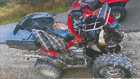 A red and black All-Terrain Vehicle, with heavy front end damage is on the gravel shoulder of a roadway, with a second red ATV seen in the background;