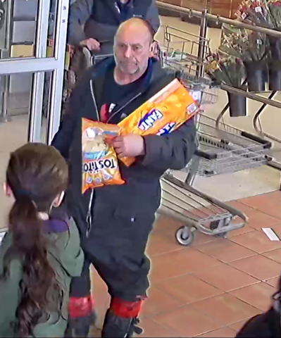 A photo of an unknown male suspect - walking through the entry/exit of a grocery store – while carrying a food item and a case of soda;