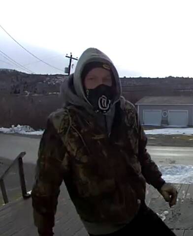 A surveillance image of a masked man – standing on a wooden deck – wearing a camouflage jacket, a grey hoodie and gloves.
