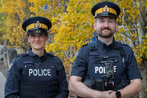 One female police officer wearing an operational RCMP uniform, including a hat, blue uniform shirt, blue uniform vest and vest with the word “police” written on it standing beside one male police officer wearing an operational RCMP uniform, including a hat, blue uniform shirt, blue uniform vest and vest with the word “police” written on it and a body-worn police camera.